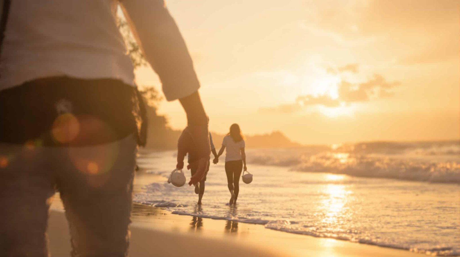 Deux personnes se tenant la main et marchant sur une plage au coucher du soleil, avec une troisième personne au premier plan.